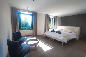 Ein modernes Hotelzimmer mit einem Bett, zwei Sessel, einem kleinen runden Tisch und großen Fenstern mit Vorhängen.