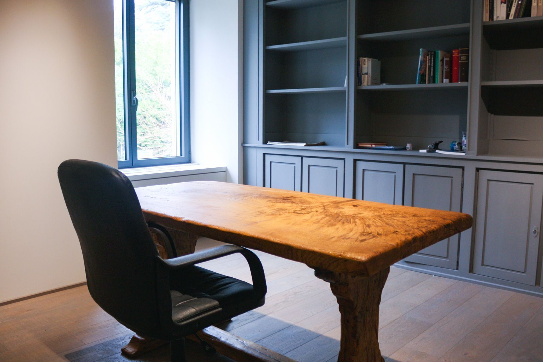 Una habitación con una mesa de madera, una silla de oficina negra, una estantería y una ventana con vistas a árboles.