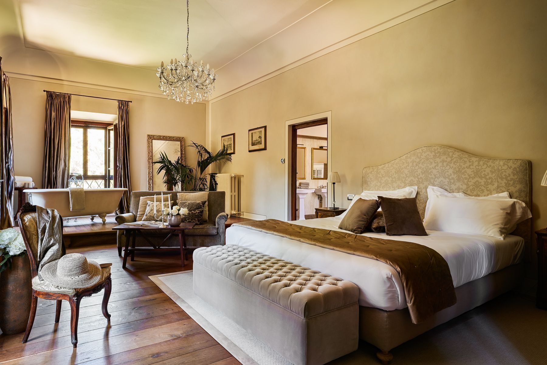 Un dormitorio con una cama grande, una zona de estar con sofá y sillón, una araña y vistas al baño.