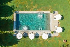 An aerial view of a rectangular swimming pool with a tiled edge, surrounded by a grassy area and white hexagonal umbrellas.