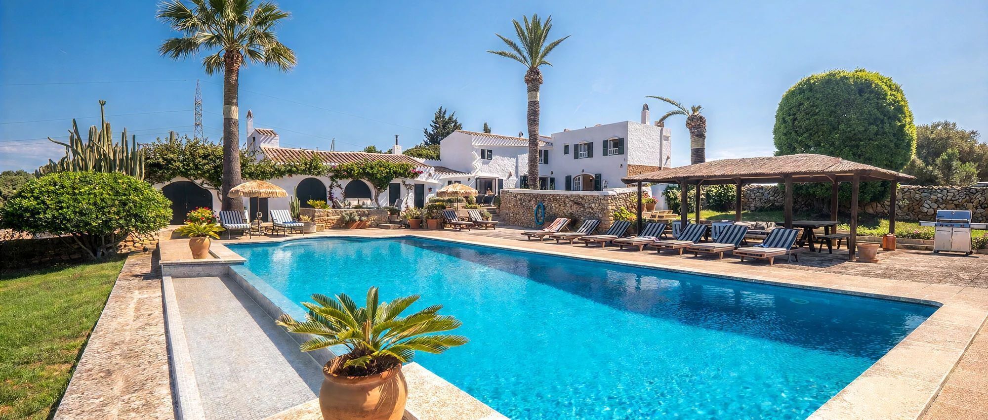 A swimming pool with lounge chairs and a gazebo, surrounded by palm trees and a stone wall.