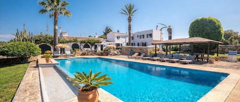 A swimming pool with lounge chairs and a gazebo, surrounded by palm trees and a stone wall.