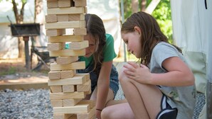 Dos personas jugando a un juego de bloques de madera al aire libre, con una tienda de campaña y árboles al fondo.