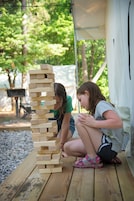 Dos personas jugando a un juego de bloques de madera al aire libre, con una tienda de campaña y árboles al fondo.