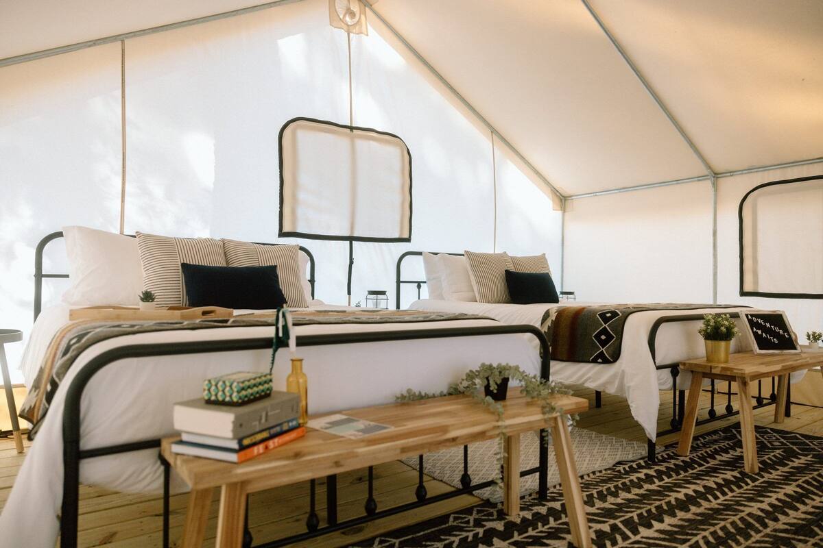 A tented accommodation with two beds, a wooden table, and a sign reading 'Adventure Awaits'.