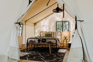 Una habitación con carpa, una cama, una mesita con un cartel, una lámpara y una ventana.