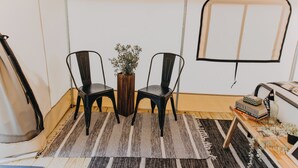 Una habitación con dos sillas negras, una mesita de madera, una planta en maceta, un tapete a rayas y una ventana con marco negro.