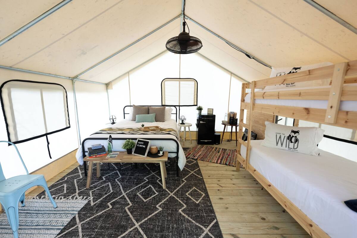 A tented accommodation with a bed, a small table with a plant, a fan, and a bunk bed.