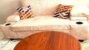 Ein beiges Sofa mit zwei gemusterten Kissen, ein runder Holztisch und eine schlichte Wand mit einem Lichtschalter.