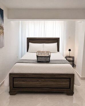 A bedroom with a large bed, a bedside table, a lamp, and a window with curtains.