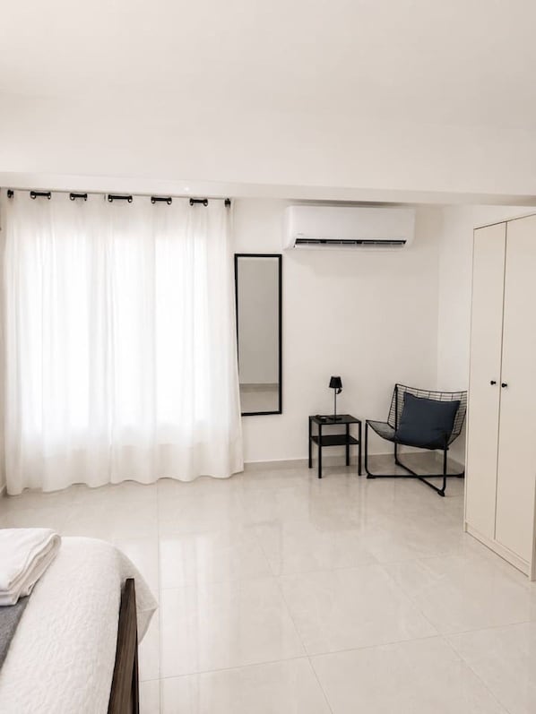 A modern hotel room with a bed, a chair, a small table, an air conditioning unit, and a wardrobe.