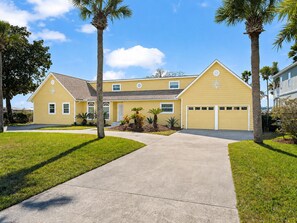 A two-story yellow house with a front driveway, palm trees, and a well-maintained lawn.
