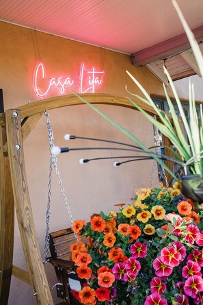 Ein hölzerner Schaukelstuhl mit einem Neon-Schild mit der Aufschrift 'Casa Lita' und einem Blumentopf voller bunter Blumen.