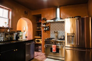 A kitchen with a stainless steel refrigerator, oven, and stove. There is a window with a view of the outside, and a shelf with various kitchen items. - Casa Lita – Colorful Historic Home in the Railyard (Santa Fe)