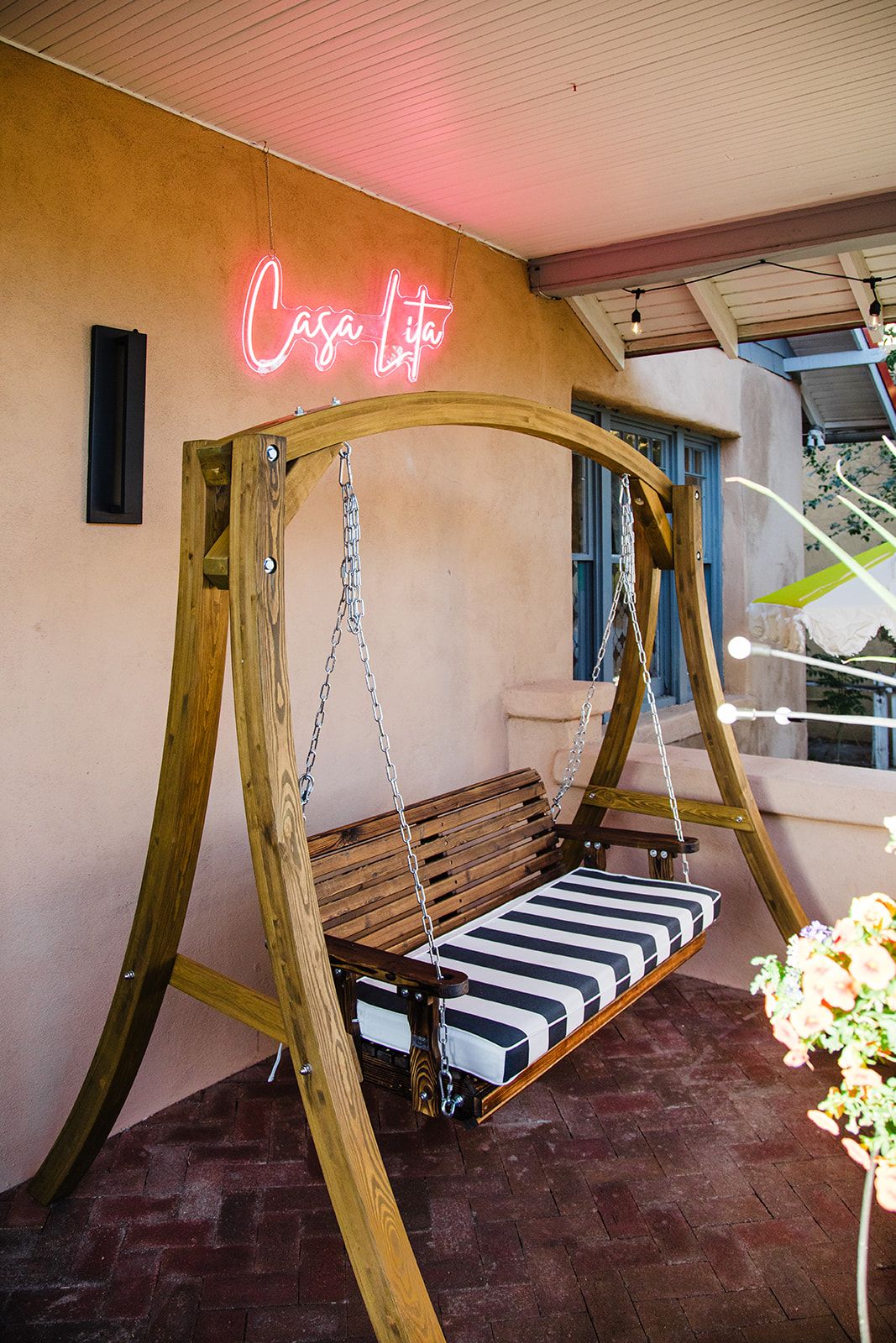 A wooden swing with a striped cushion, a neon sign reading 'Casa Lita,' and a brick floor.