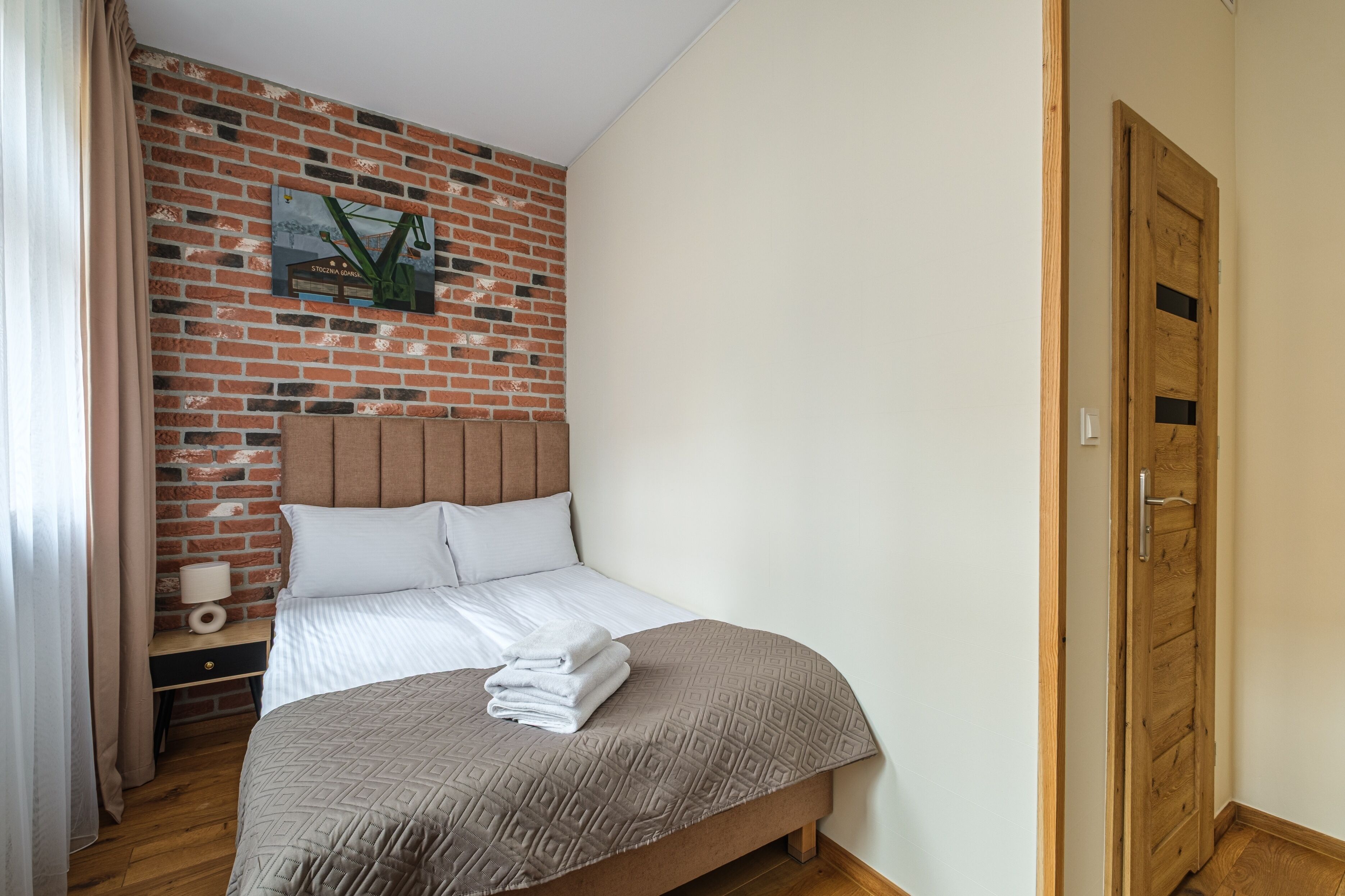 A hotel room with a brick wall, a wooden door, a bed with a headboard, a nightstand with a lamp, and a wall-mounted picture.