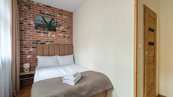 A hotel room with a brick wall, a wooden door, a bed with a headboard, a nightstand with a lamp, and a wall-mounted picture.