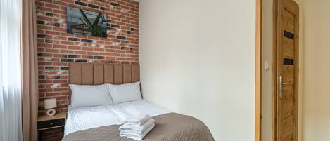 A hotel room with a brick wall, a wooden door, a bed with a headboard, a nightstand with a lamp, and a wall-mounted picture.