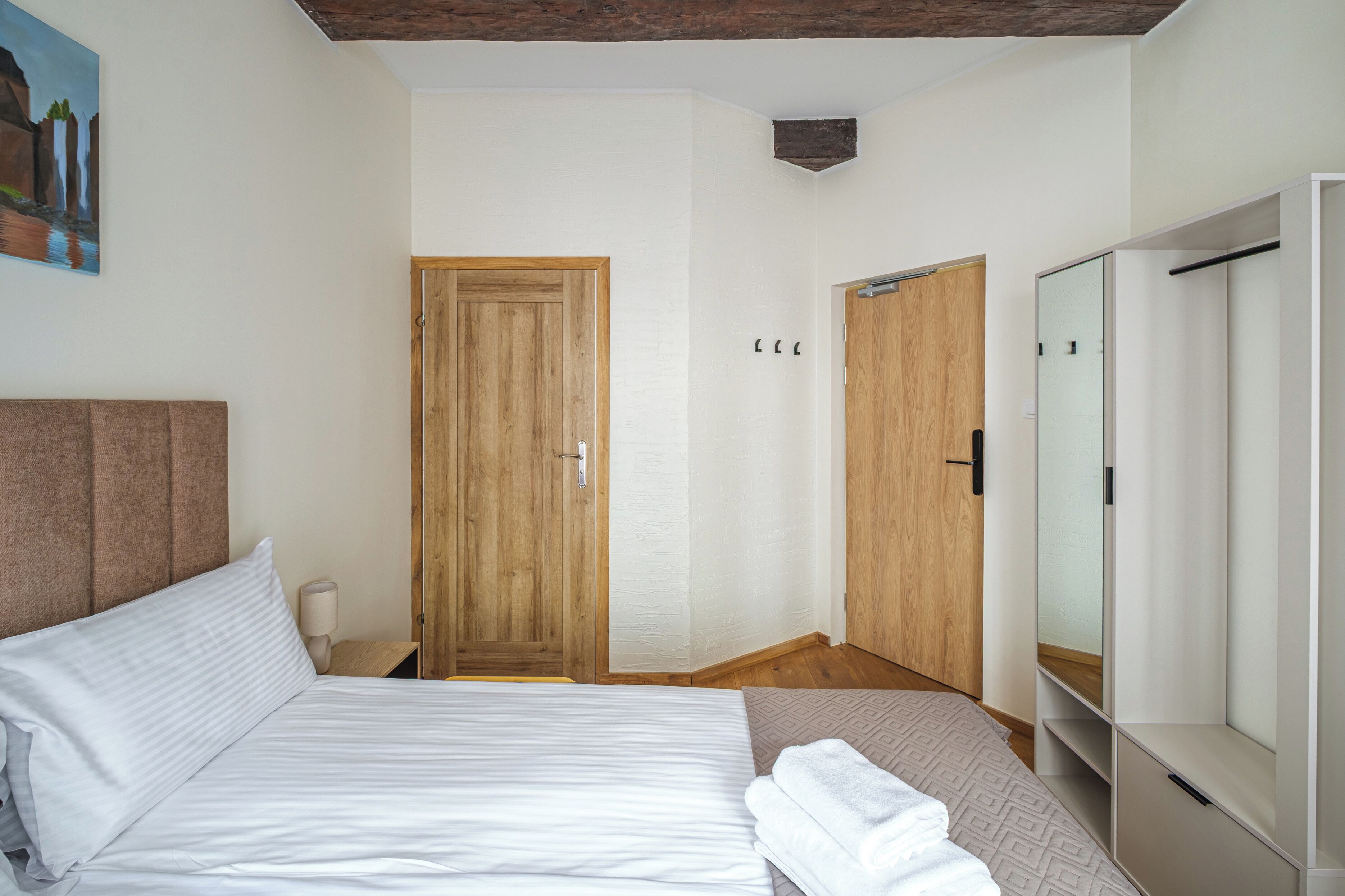 A hotel room with a bed, a nightstand, a lamp, a wooden door, a wardrobe, and a painting on the wall.