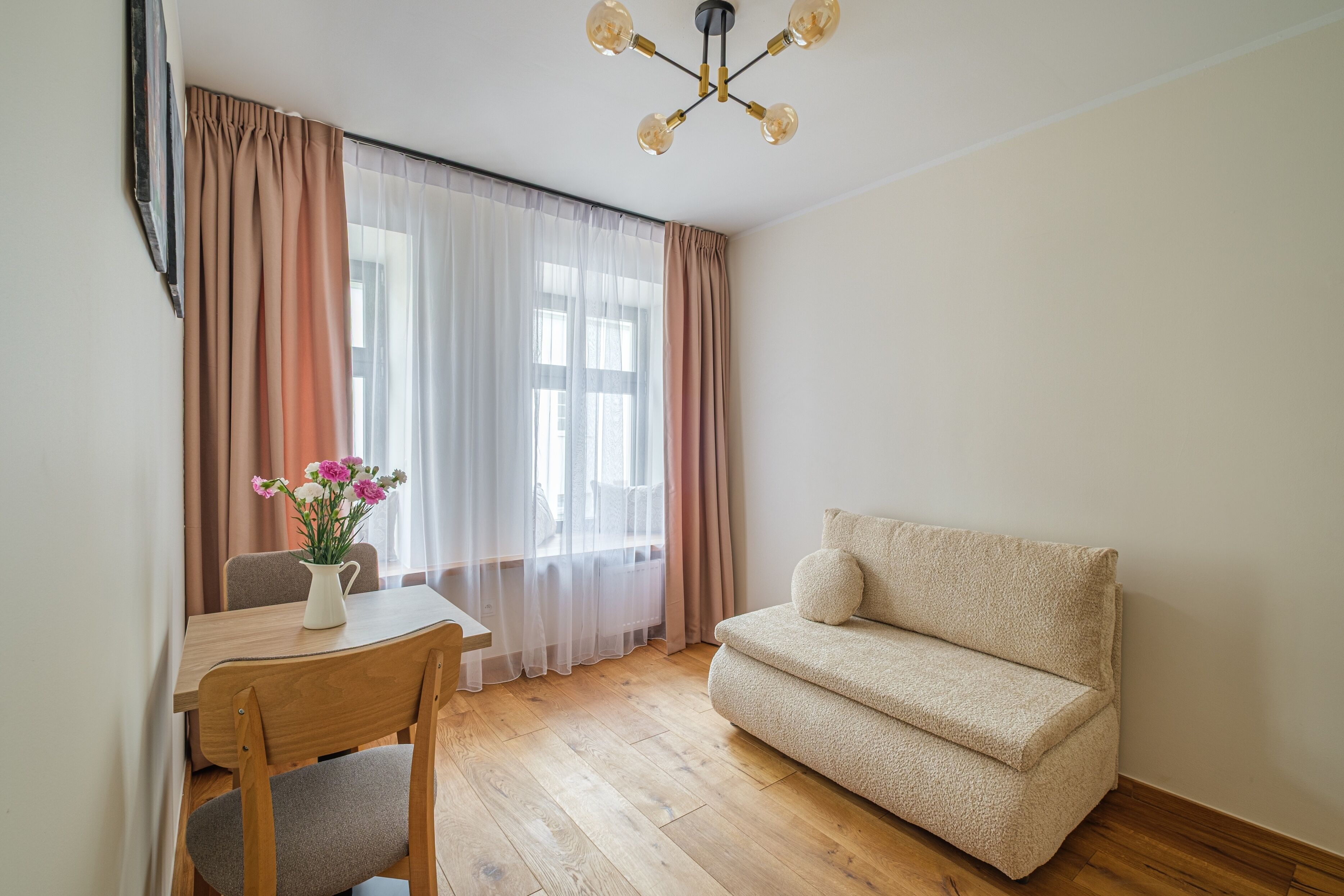 A room with a wooden floor, a beige sofa, a wooden table with a vase of flowers, and sheer curtains.