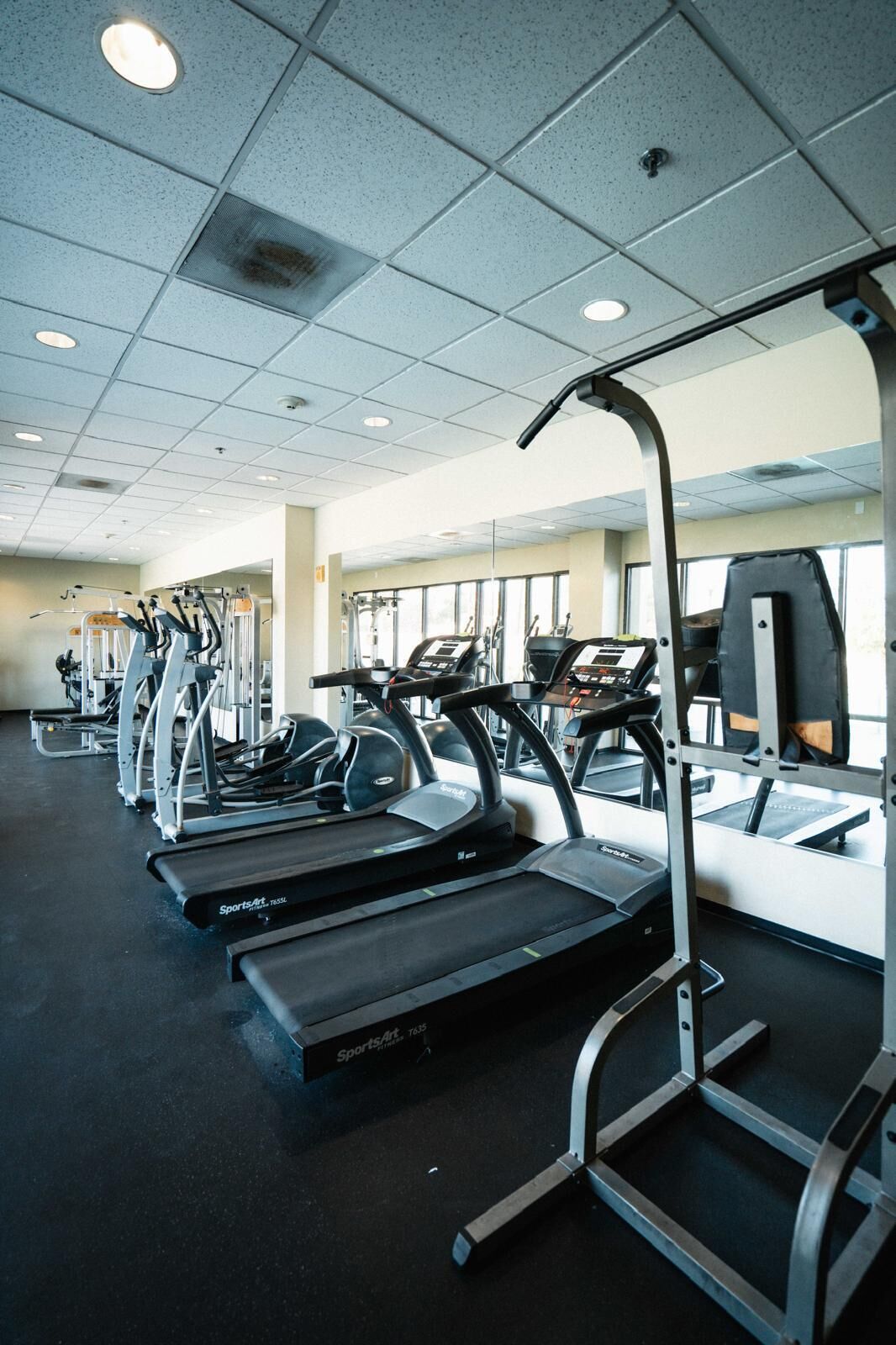 A gym with multiple treadmills and exercise machines, large windows, and a tiled ceiling.