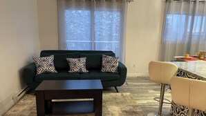 A living room with a dark sofa, patterned cushions, a coffee table, a bar stool, and a view through sheer curtains. - Spacious newly renovated two bedroom apartment (Sault Ste. Marie)