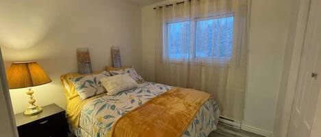Un dormitorio con cama, mesita de noche, lámpara, ventana con cortinas y puerta.