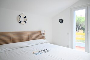 Um quarto com cabeceira de madeira, cama com roupa de cama branca, decoração com boia salva-vidas, leme de navio e vista para o verde lá fora.