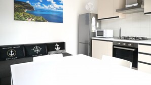 Eine moderne Küche mit Herd, Mikrowelle und Kühlschrank. Ein Esstisch mit weißem Tischlaken und schwarzen Stühlen steht dort. Ein Gemälde einer Küstenlandschaft hängt an der Wand.