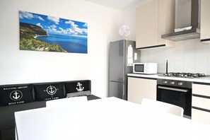 Eine moderne Küche mit Herd, Mikrowelle und Kühlschrank. Ein Esstisch mit weißem Tischlaken und schwarzen Stühlen steht dort. Ein Gemälde einer Küstenlandschaft hängt an der Wand.