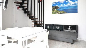 Ein modernes Wohnzimmer mit einem weißen Esstisch, einem grauen Sofa mit nautisch gestalteten Kissen, einer schwarzen Holztreppe und einem großen, atmosphärischen Wandbild.