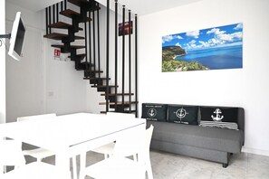 Uma sala de estar moderna com uma mesa de jantar branca, um sofá cinza com almofadas de temática náutica, uma escada preta e de madeira, e uma grande obra de arte com paisagem.