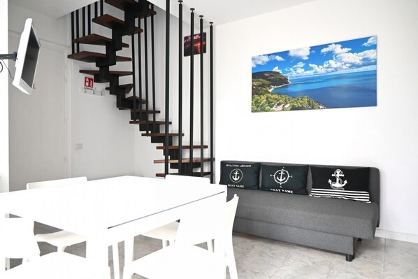 Ein modernes Wohnzimmer mit einem weißen Esstisch, einem grauen Sofa mit nautisch gestalteten Kissen, einer schwarzen Holztreppe und einem großen, atmosphärischen Wandbild.