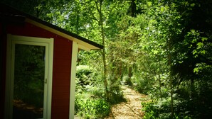 Um edifício vermelho com porta e janela, cercado por folhagem verde densa e um caminho arborizado.