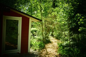 Un edificio rojo con una puerta y una ventana, rodeado de follaje verde exuberante y un sendero arbolado.