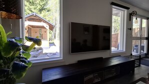 Uma sala de estar moderna com uma grande TV de tela plana na parede, um rack de madeira escura e grandes janelas com vista para uma cabana de madeira e árvores.
