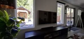 Una sala de estar moderna con un televisor de pantalla plana grande montado en la pared, un mueble de entretenimiento de madera oscura y ventanas amplias que ofrecen vistas a una cabaña de madera y árboles.