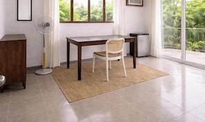 Habitació amb escriptori de fusta, cadira blanca, ventilador vertical i vistes a la verdor a través d'una finestra gran.