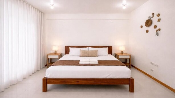 A bedroom with a wooden bed, white bedding, two bedside tables with lamps, and a large window with white curtains. - ISLO APT (General Luna)