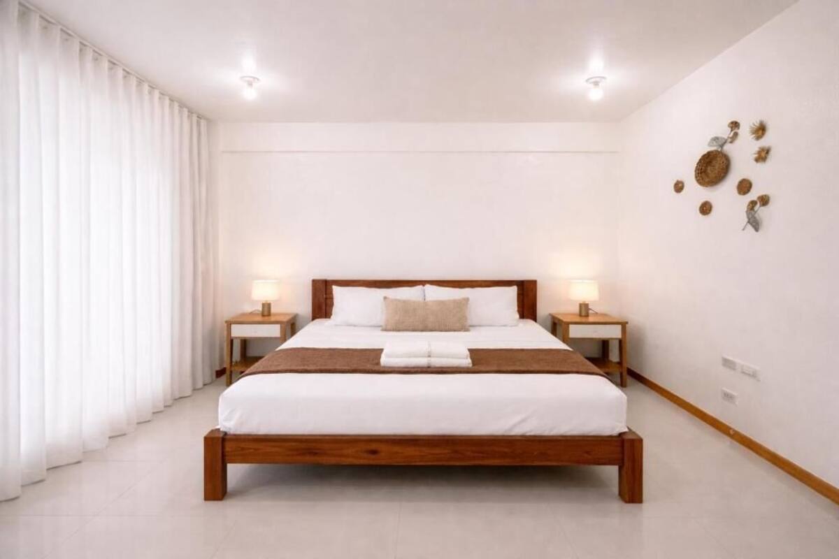 A bedroom with a wooden bed, white bedding, two bedside tables with lamps, and a large window with white curtains.