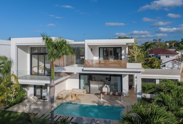 Ein modernes, zweistöckiges Haus mit Swimmingpool, Sitzgelegenheiten im Freien und üppiger Bepflanzung.