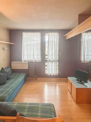 Ein Zimmer mit einer Couch, einem Fernseher, einem Schreibtisch und zwei Fenstern mit Vorhängen.