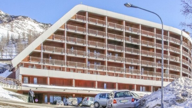 Ein mehrstöckiges Gebäude mit bogenförmiger Fassade, ein Parkplatz mit Autos und eine Berglandschaft im Hintergrund.