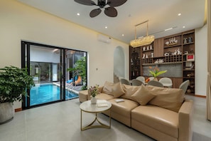 Ein modernes Wohnzimmer mit einer Couch, einem Couchtisch, einer Kristallleuchter, Blick auf einen Pool und einem Bücherregal.