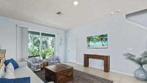 Een woonkamer met een bank, salontafel en gestreepte stoelen. Er is een open haard, een schilderij van een strand en een deur die naar buiten leidt.