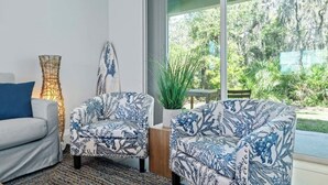Een moderne woonkamer met een bank, twee fauteuils met een patroon, een hoge lamp en uitzicht op groen buiten.