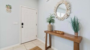 Een hal met een witte deur, een houten console tafel met een spiegel, en twee potplanten.