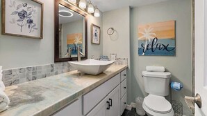 Et badeværelse med en hvid vask, toilet og et spejl med lys. Væggene er dekoreret med billeder, og der er en håndklædestang.