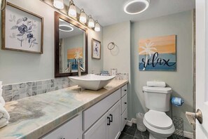 A bathroom with a white sink, a toilet, and a mirror with lights. There are framed pictures on the wall and a towel rack. - Sunrise Beachside Oasis & Pool (Jacksonville Beach)