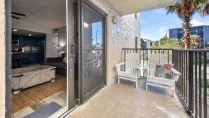 A balcony with a sliding glass door leading to a modern kitchen, a white chair with a floral cushion, and a palm tree in the background.